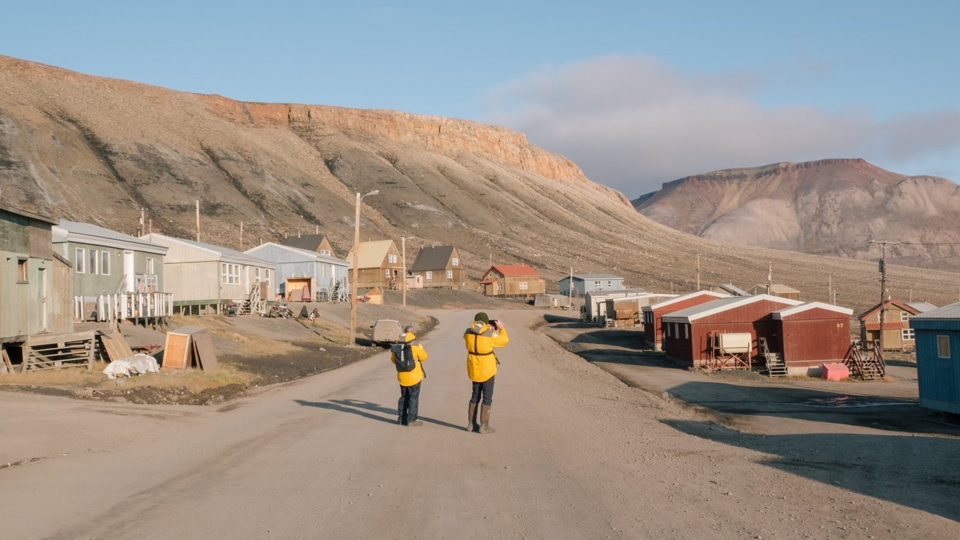 Two expedition guests in yellow Quark parkas walking down the main road of Arctic Bay, Nunavut, beneath the hills.
