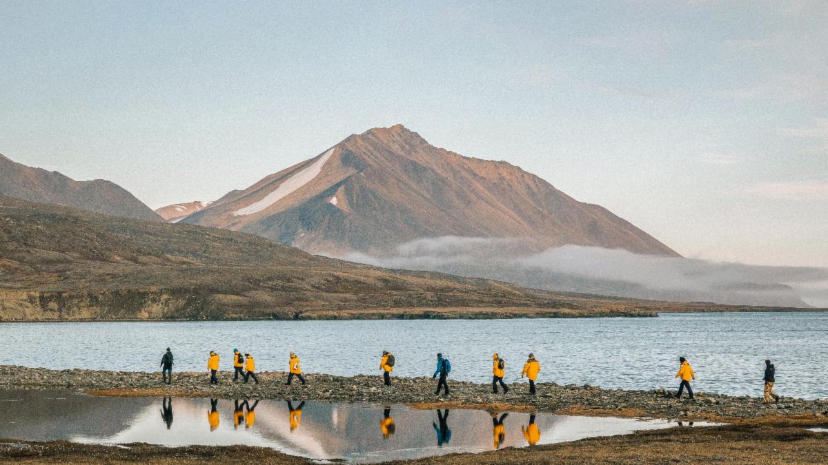 Expedition guests in yellow parkas walking single file along the tundra shoreline beneath an Arctic mountain.