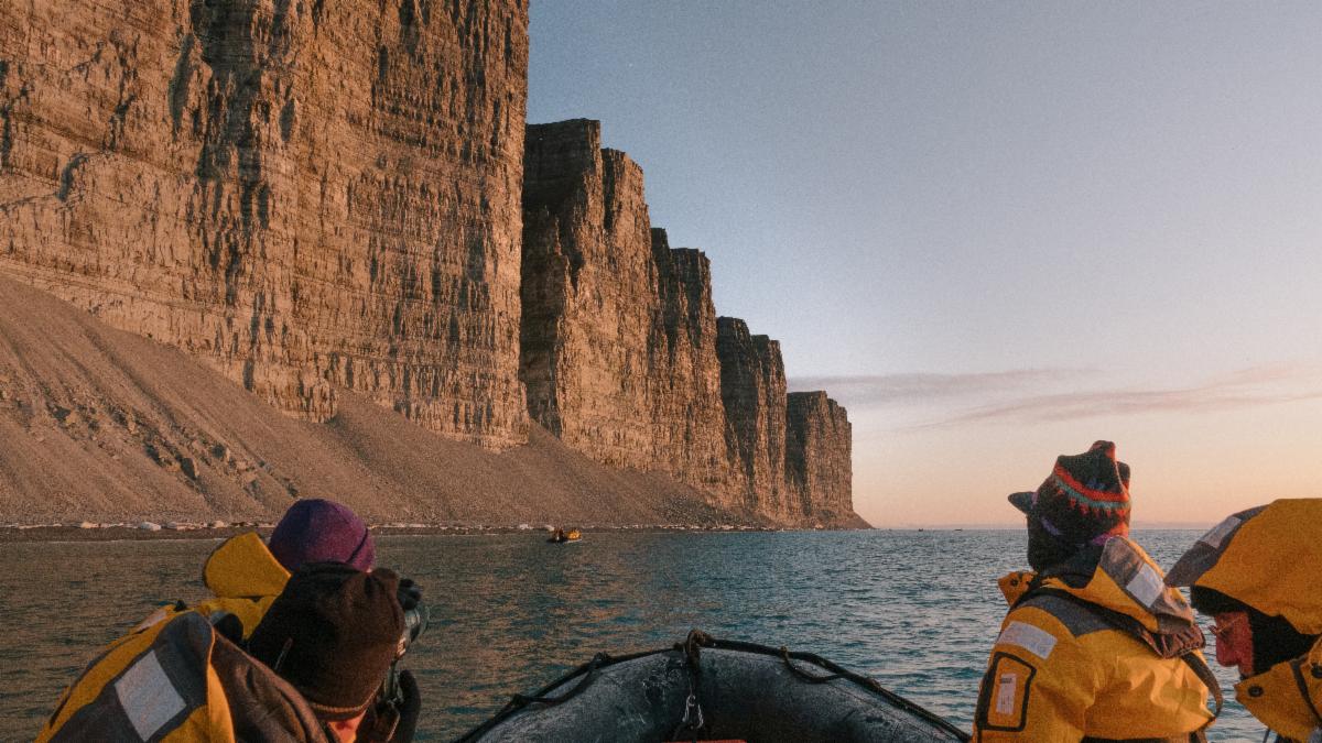 Guests in a Zodiac beneath sunlit Arctic sea cliffs.